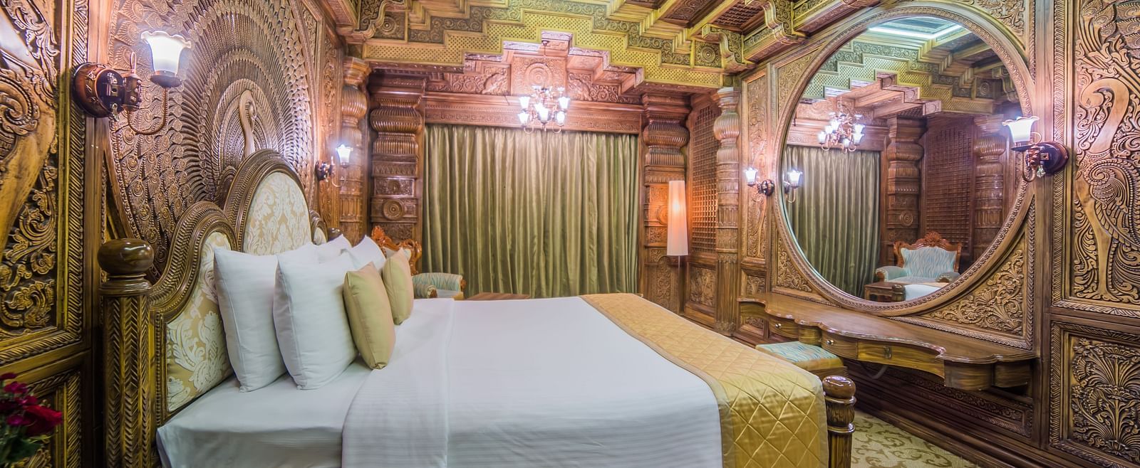 An intricately carved room with traditional motifs, a plush bed, and rich wooden decor A grand hotel suite with an opulent red and white decor, large bed, and seating area, reflecting a traditional style at Sitara Luxury Hotel in Hyderabad near Ramoji Film