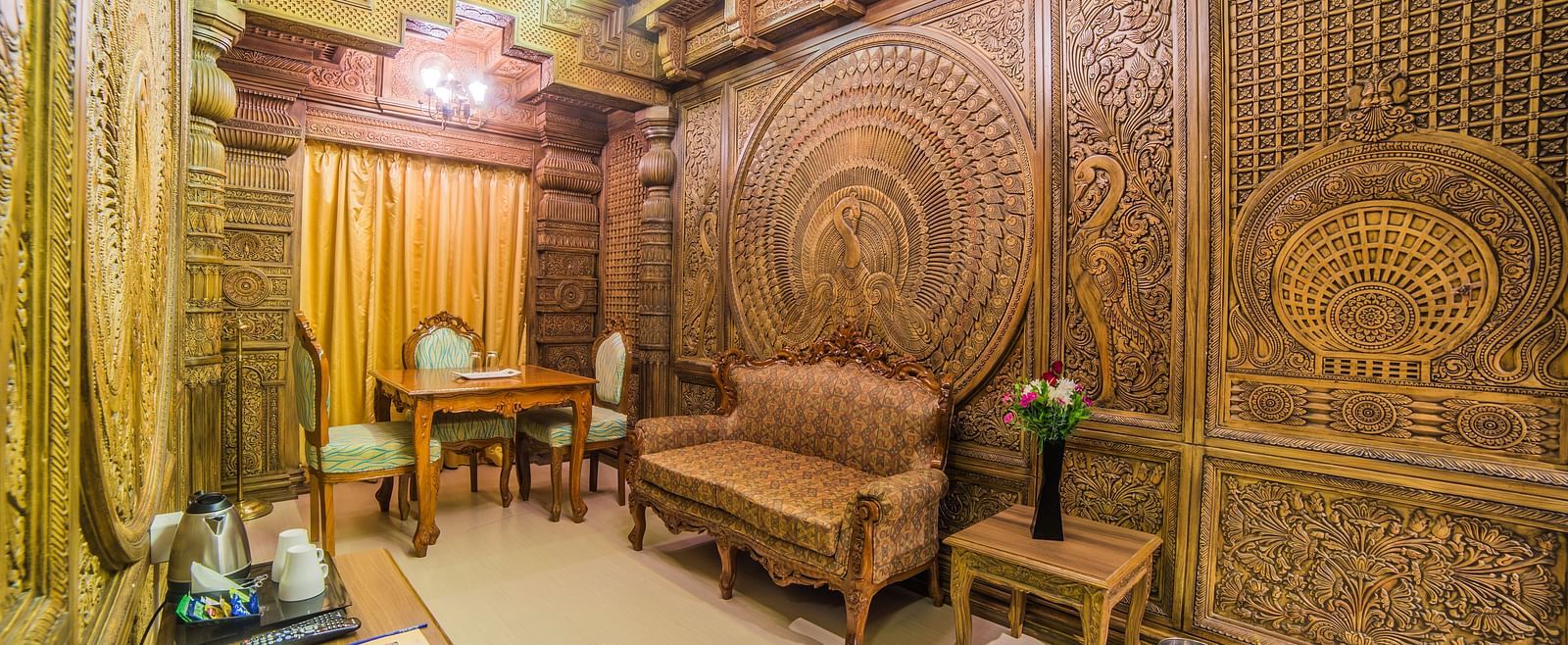 A spacious seating area with ornate wood carvings and classic furniture A grand hotel suite with an opulent red and white decor, large bed, and seating area, reflecting a traditional style at Sitara Luxury Hotel in Hyderabad near Ramoji Film City.