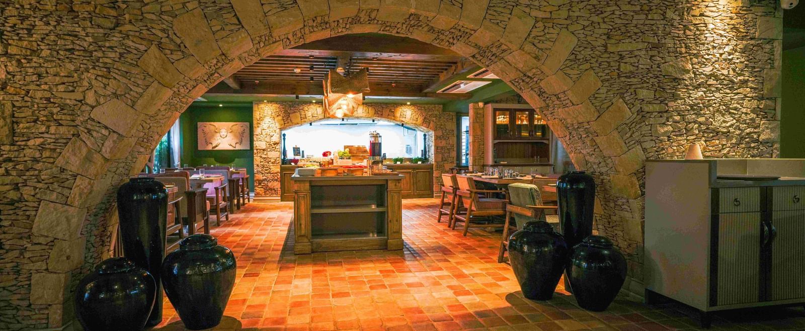 A view into Mila Restaurant at The Evren, Vagator, taken through a rustic stone archway. The restaurant has a tiled floor, a central buffet area, and various dining tables, creating a warm and inviting atmosphere.