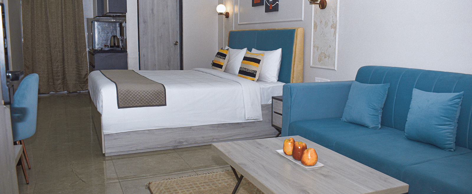 A hotel room with a bed, a blue sofa, a light wood coffee table with fruit, a kitchenette area, and wall-mounted light fixtures - Urban Suites by BluSalzz, Jaipur