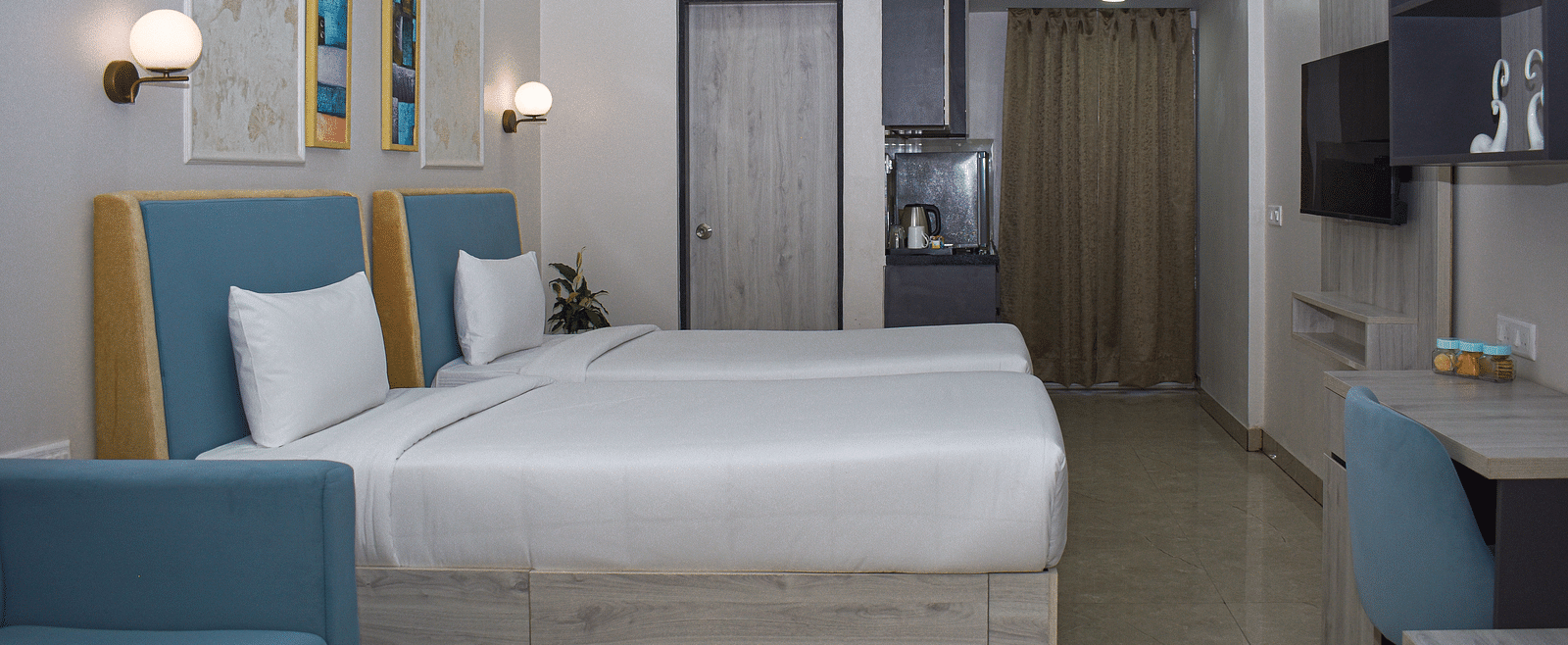 A hotel room with two twin beds, a blue sofa, a coffee table, a desk and chair, a wall-mounted TV, and a kitchenette area - Urban Suites by BluSalzz, Jaipur