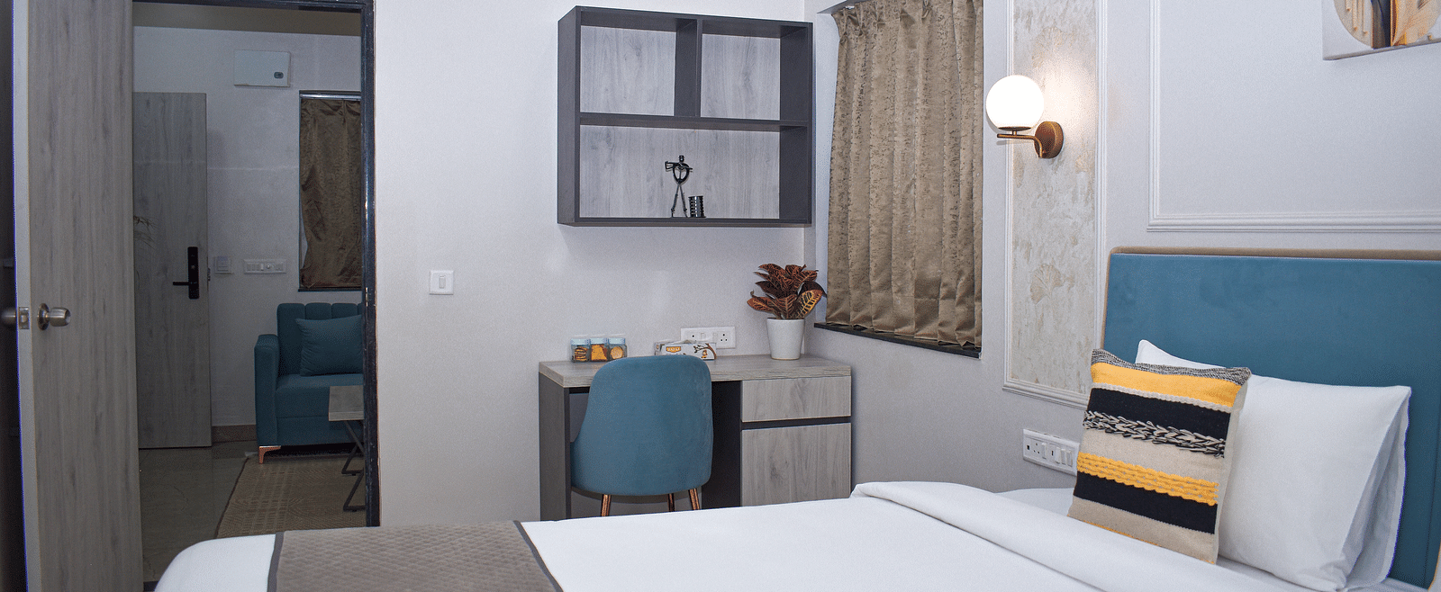 A hotel bedroom with a bed, a desk and chair, a wall cabinet, a window with curtains, and a doorway leading to another room - Urban Suites by BluSalzz, Jaipur