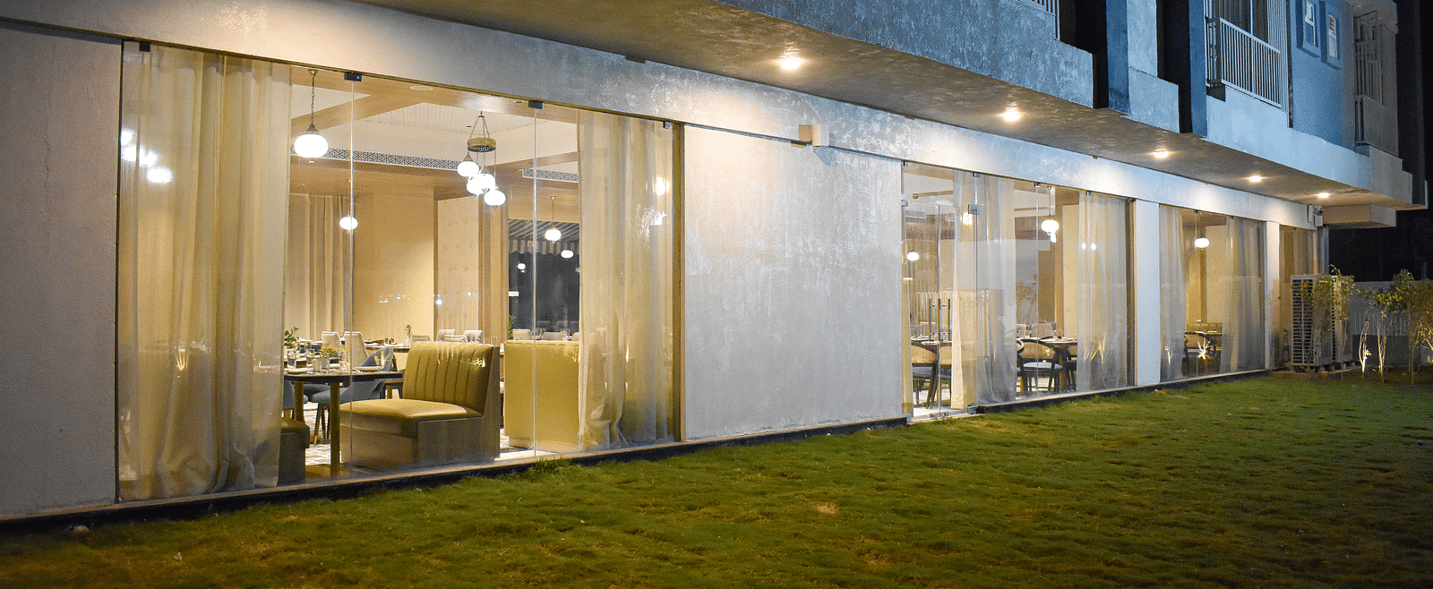 Exterior view of a building with a glass-walled restaurant, sheer curtains, hanging lights, and a grassy area in front - Urban Suites by BluSalzz, Jaipur