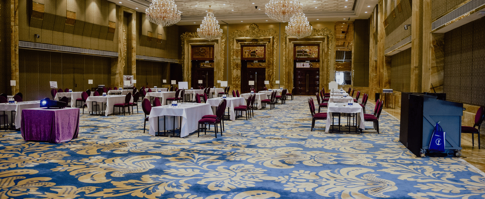 A spacious banquet hall with ornate chandeliers, patterned carpet, and tables set up for an event or conference.