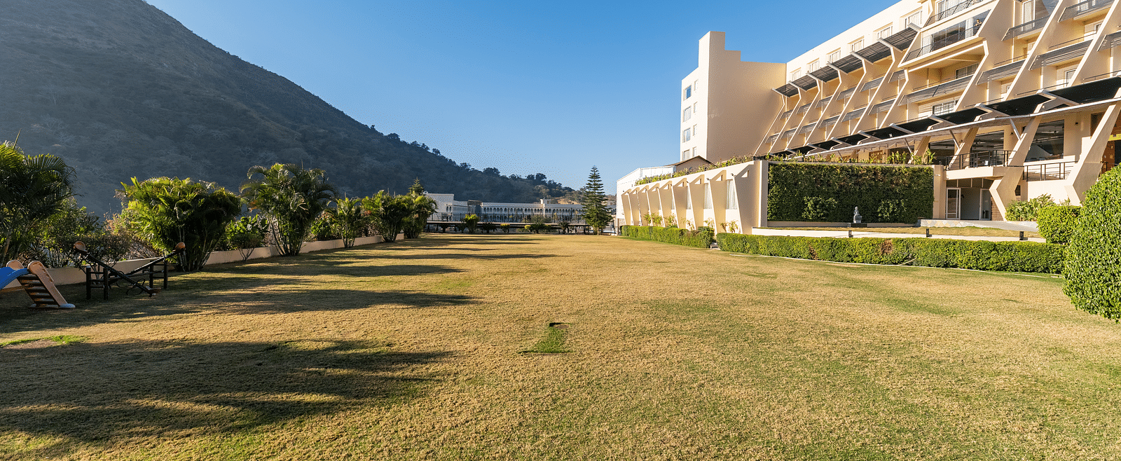 A wide shot of a building with a mountain backdrop in a green field - Via Lakhela Resort & Spa