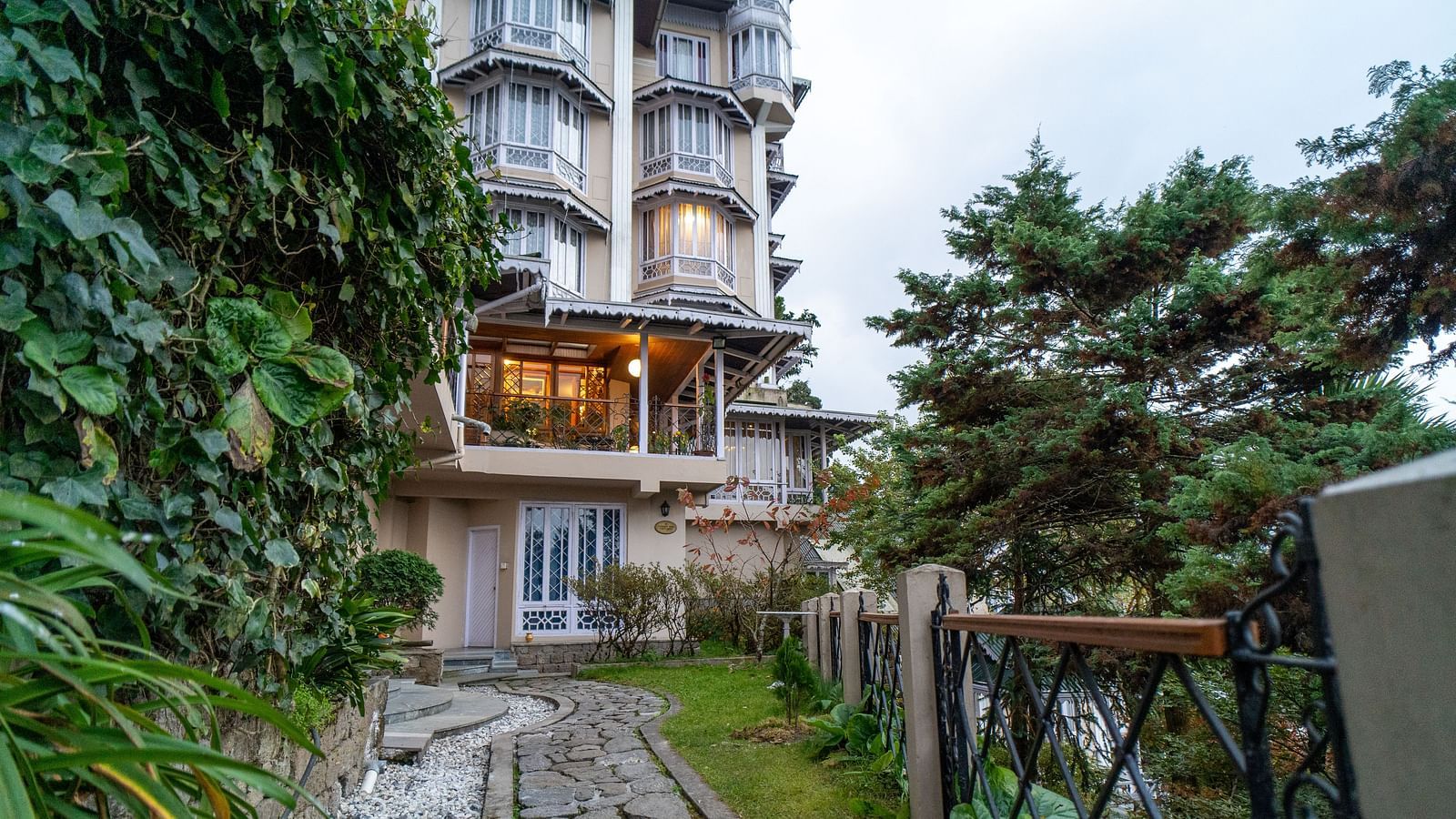 A narrow pathway leading to the charming entrance of Cedar Inn, Darjeeling, lined with greenery and wrought iron fencing.