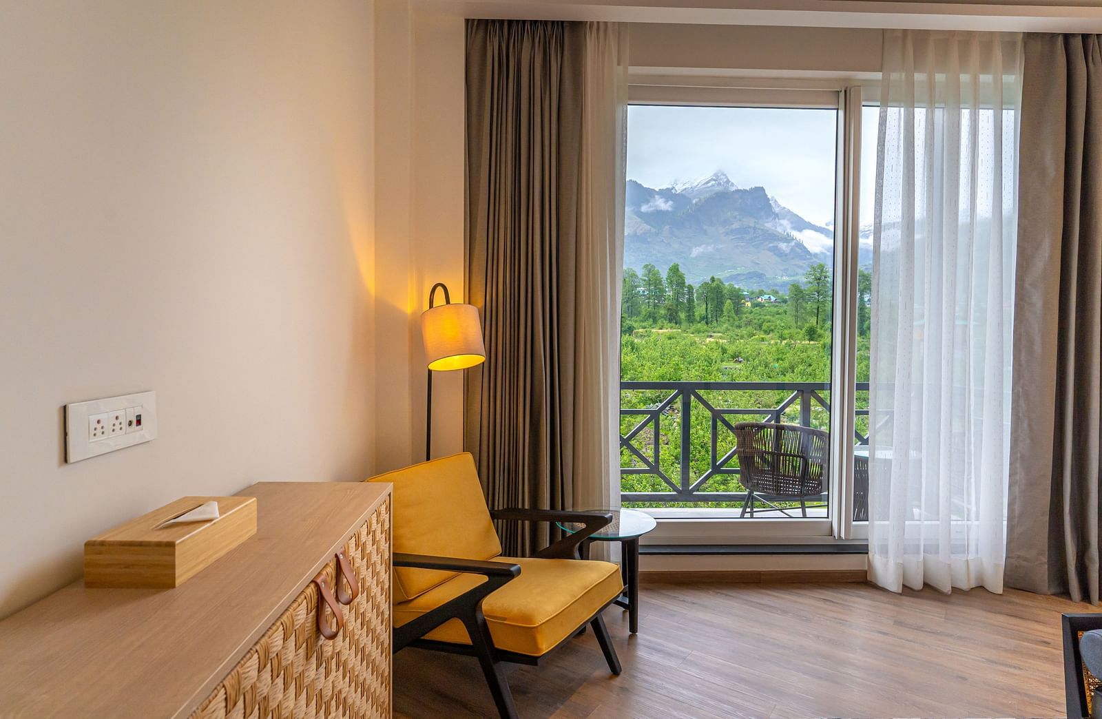 Wooden table placed along the wall with a cushioned chair, lamp beside it, and a glass door leading to the balcony at Nivasana Manali.