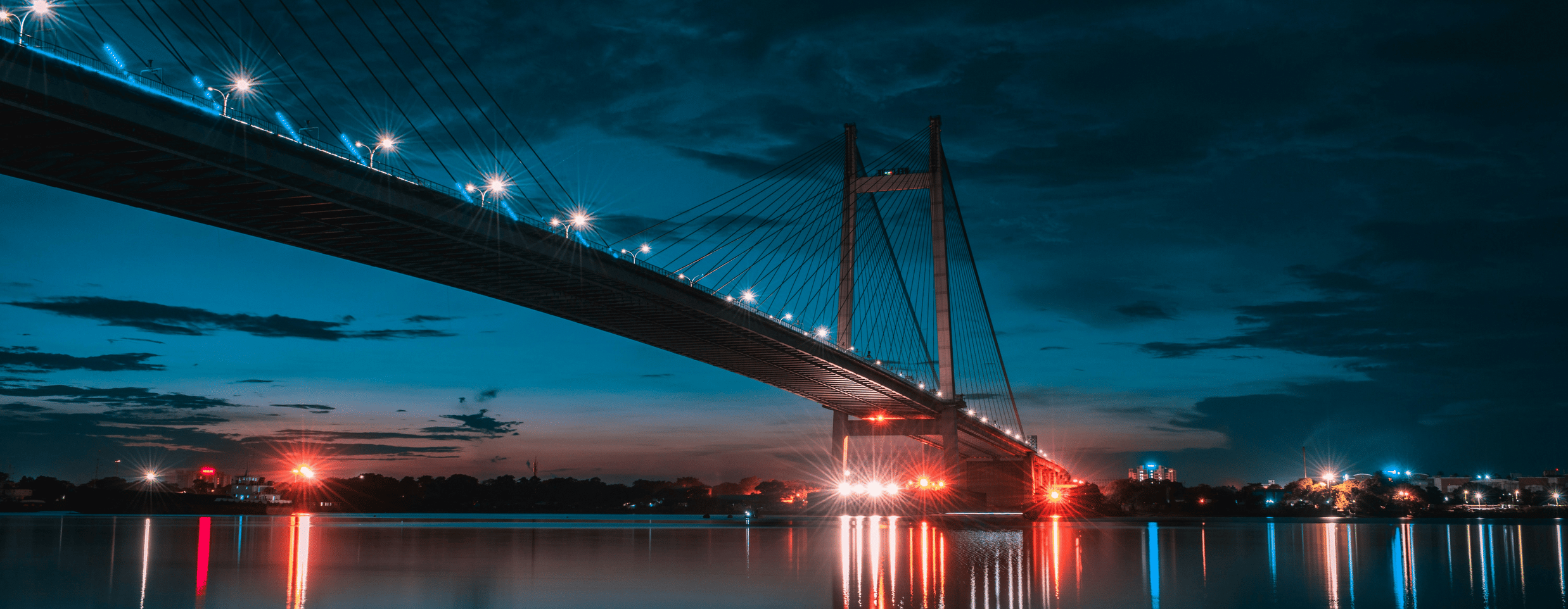 Suspension bridge spanning the Hooghly River with reflections on the water at night, at Howrah Bridge, Kolkata.
