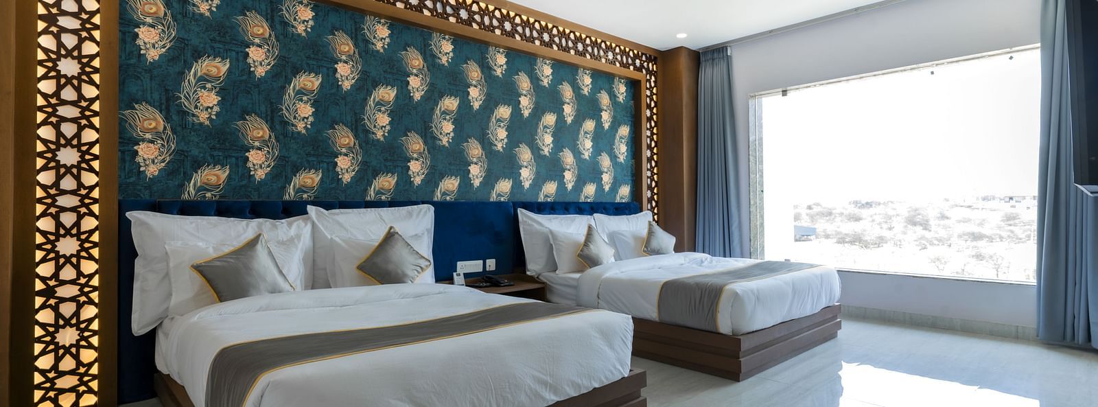A bedroom at the Bamboo Saa Mulberry Resort featuring two twin beds with white linens and grey throws. A decorative wall panel with a blue and gold pattern is above the beds and a large windows is visible.