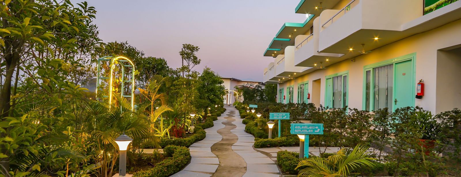 A pathway illuminated at dusk winds through the grounds of Vedikant Resorts The Mallard Corbett, showcasing the buildings.
