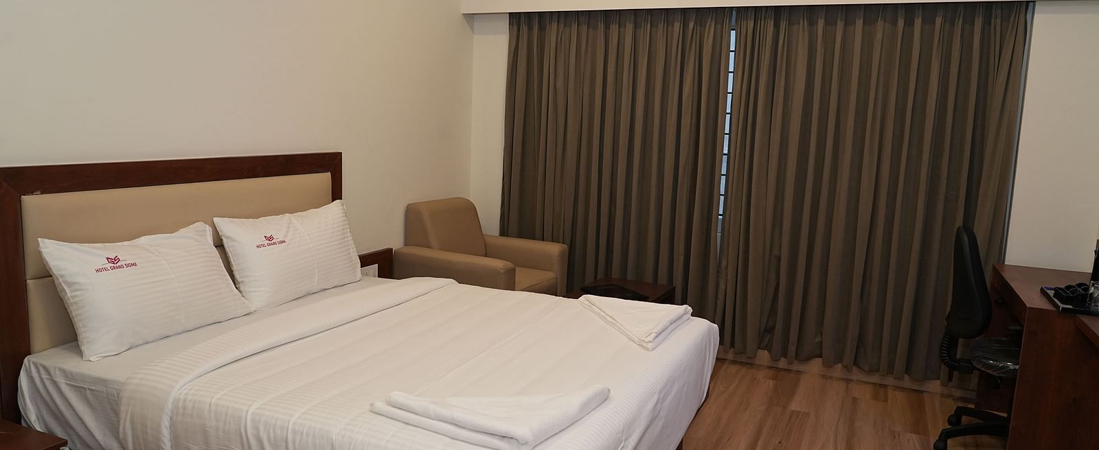 corner view image of the double bed with a huge window in the corner covered with brown curtains - Hotel Grand Sigma
