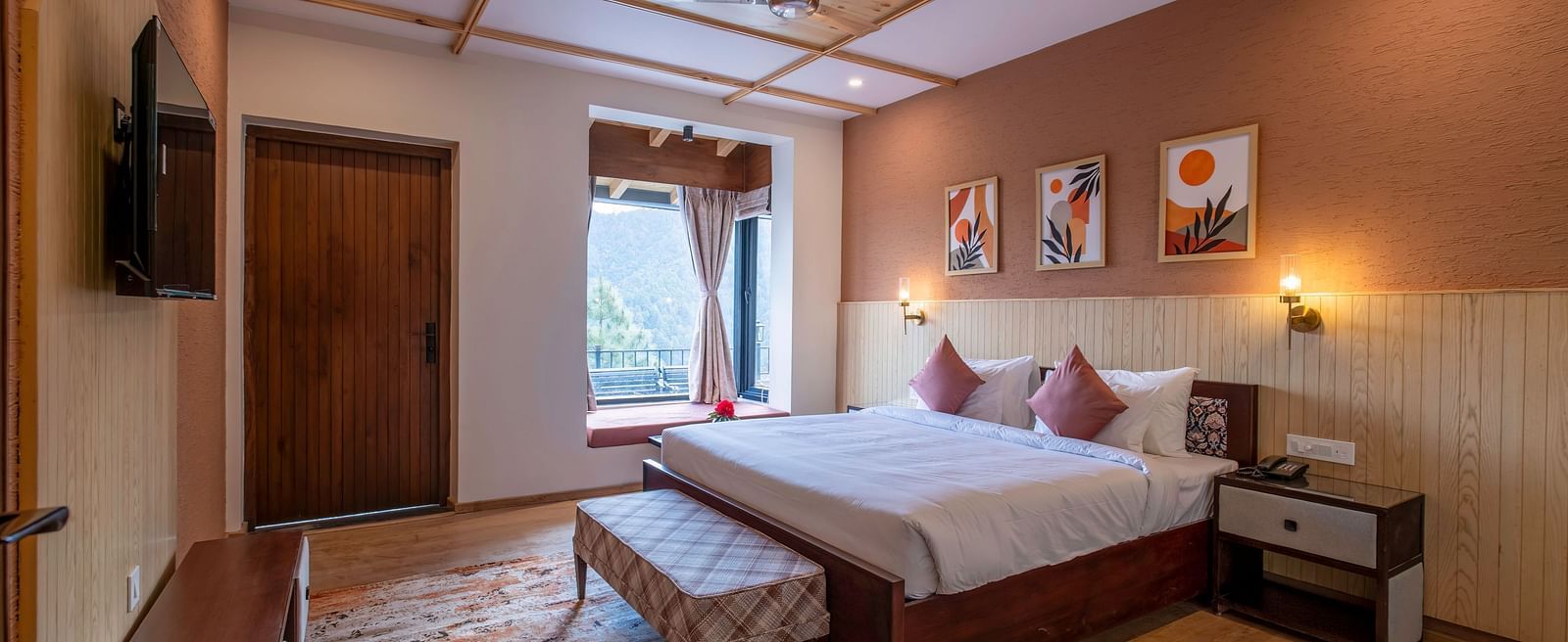 Spacious room with a large bed, bedside table, wall-mounted light above the headboard, a TV with a desk below, and windows and a door to the side at The Chamomile Estate.