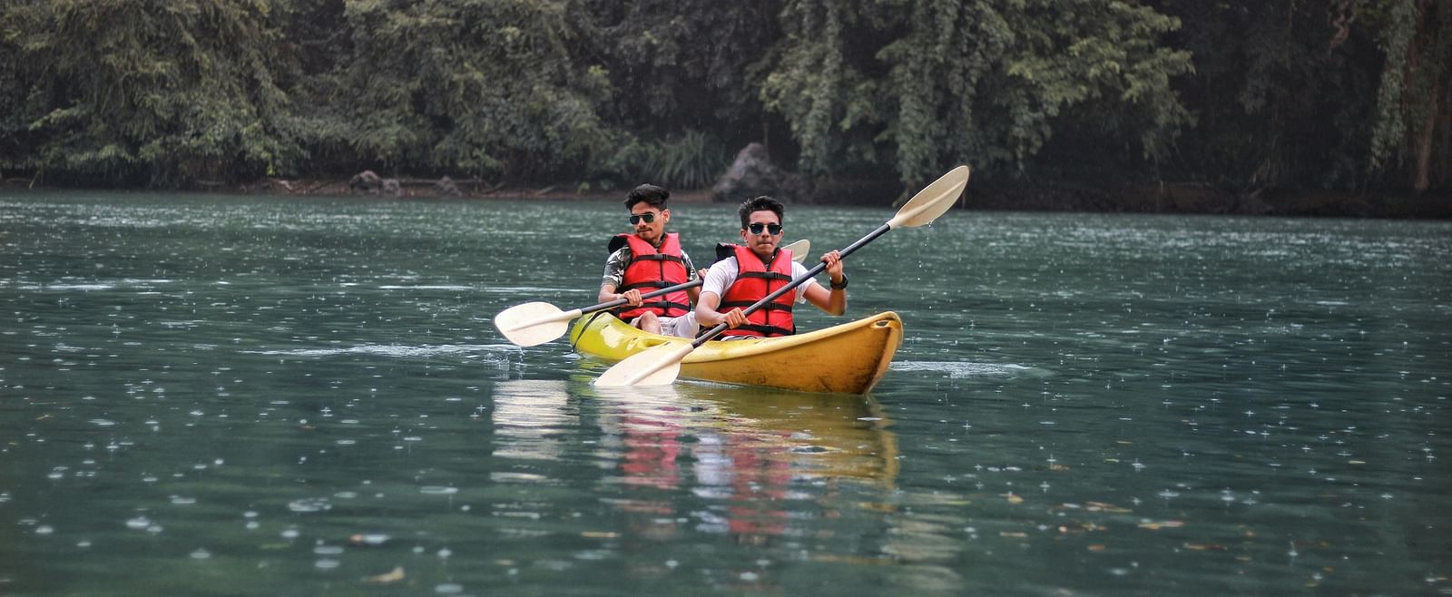 Two people kayaking on a dark water body surrounded by dense green forest.