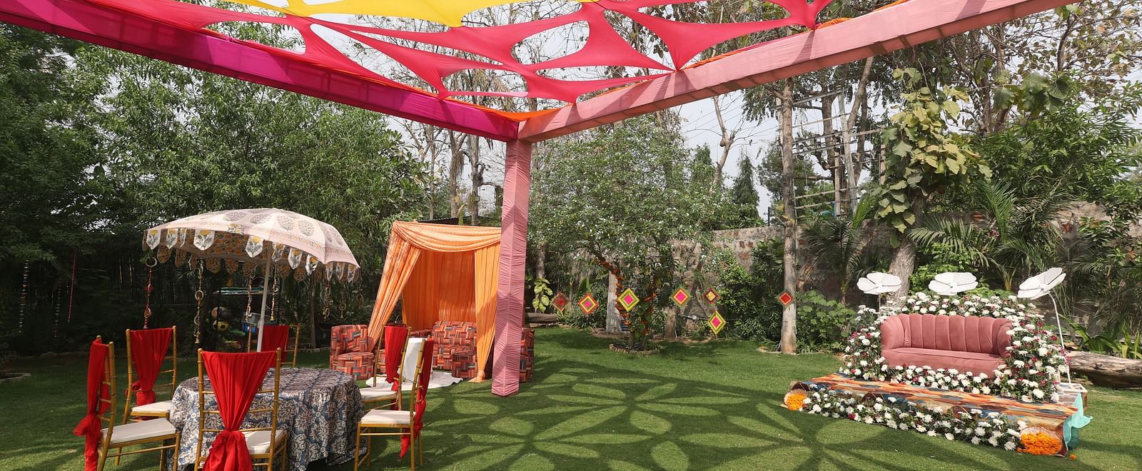 Outdoor wedding setup at Tisya Farms, Gurgaon with red chairs, and floral seating area under colourful drapes.