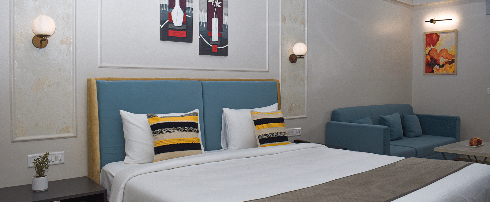 A hotel room with a bed, a blue sofa, a small table, wall-mounted light fixtures, and framed artworks - Urban Suites by BluSalzz, Jaipur