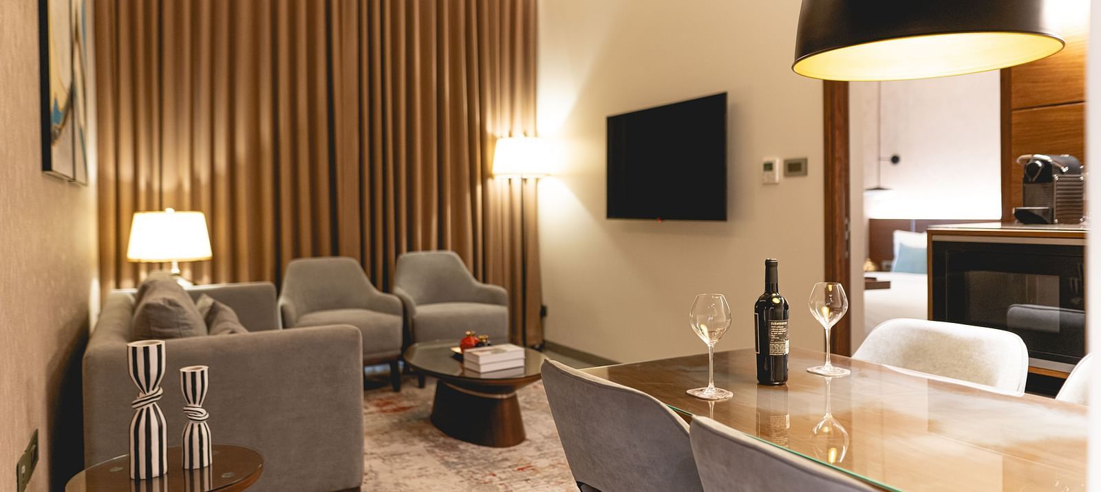 Modern living room at KertelSuites with neutral tones, featuring a sofa, armchairs, TV on the wall, floor and table lamps, and a dining table with chairs; warm lighting and vertical wooden slats on one wall