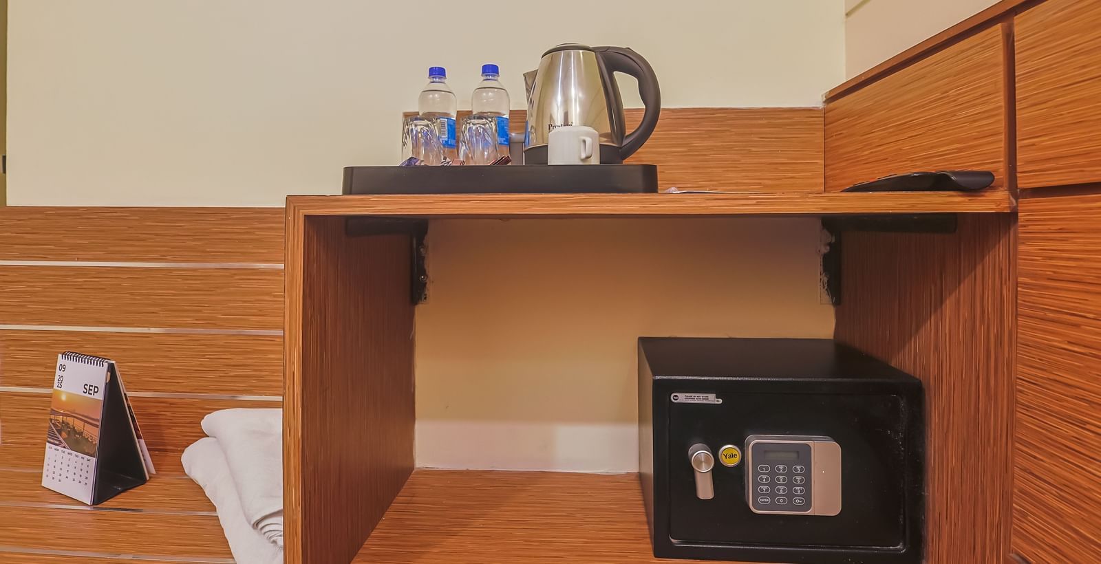 Room corner with wooden shelf, kettle, water bottles, glasses and digital safe at Max Hotel Prayagraj.