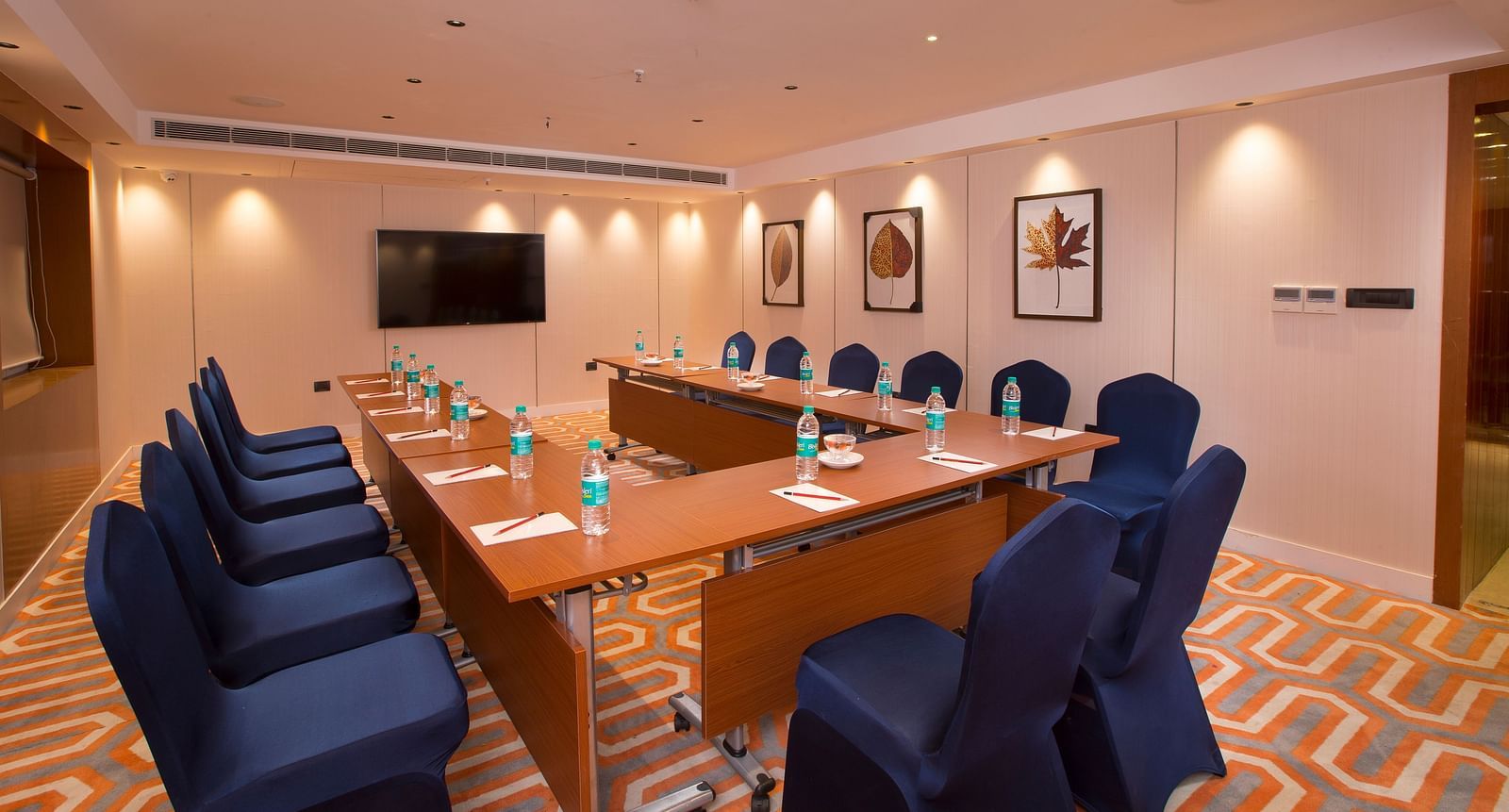 A formal boardroom with blue chairs around a long table, a flat-screen TV on the wall and framed leaf prints - Hotel Southend by TGI - Bommasandra, Bangalore