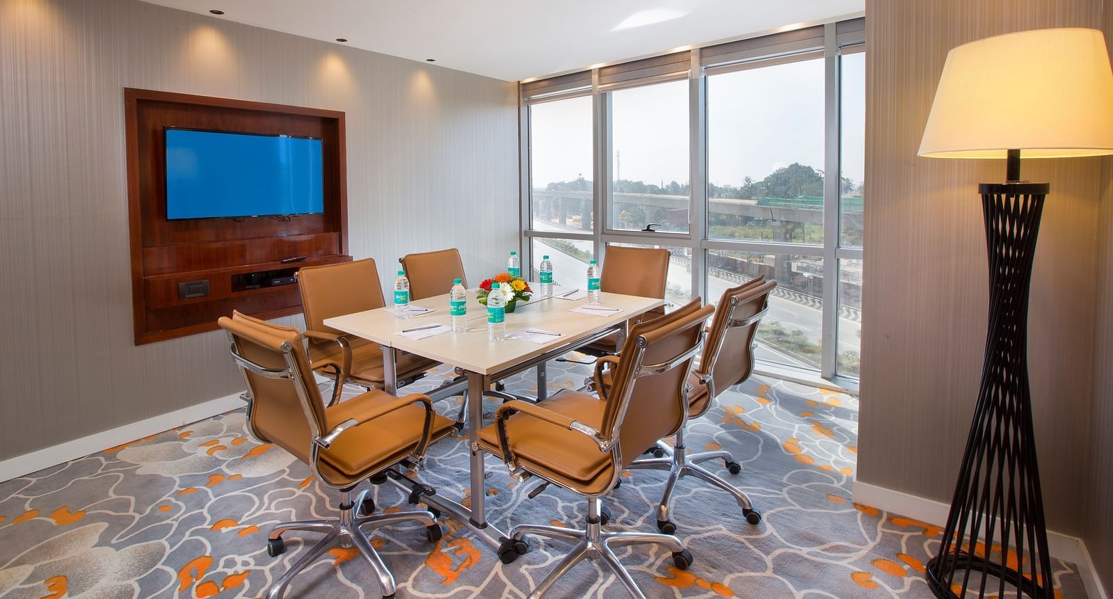 A small meeting room with a round table, tan leather chairs and a TV, with a large window and a floor lamp - Hotel Southend by TGI - Bommasandra, Bangalore