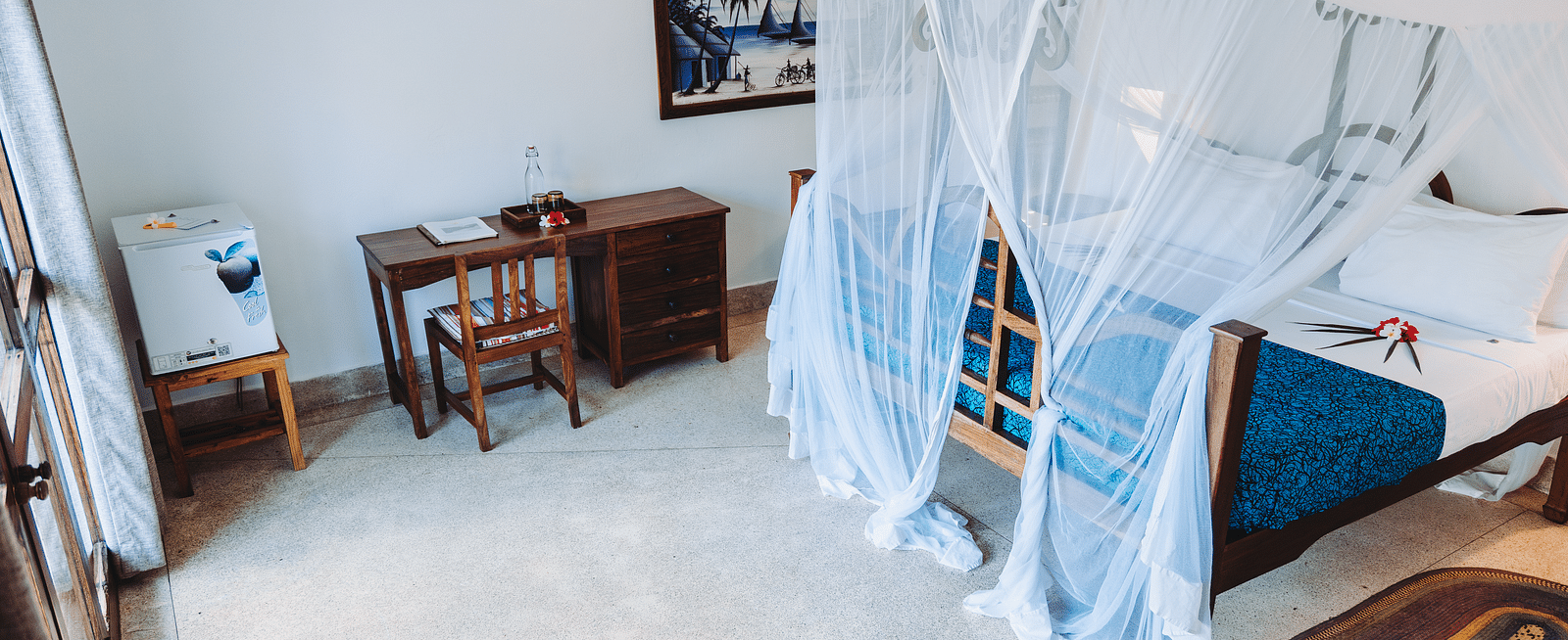 Interior view of a bedroom with a wooden four-poster bed with a mosquito net, a bedspread, a desk with a chair, a framed picture on the wall, and a small refrigerator - Shooting Star Boutique Hotel.