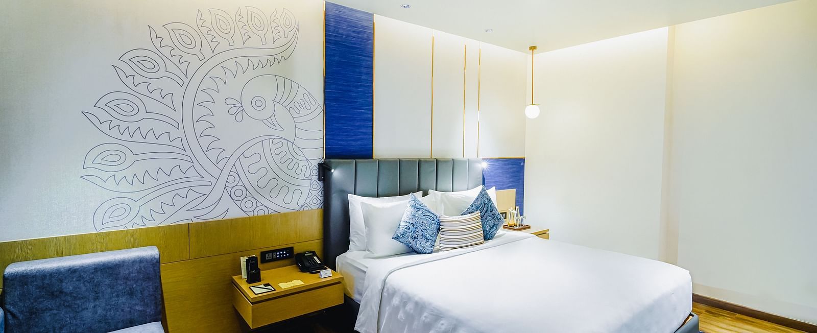 An image of our Deluxe Suite in Madurai with a peacock wall paper, a large bed, a night stand and a blue couch