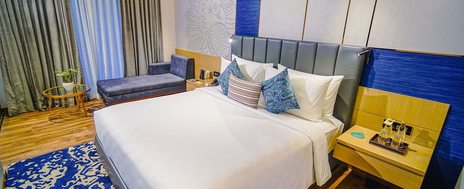 An image of a hotel room in Madurai with a large bed, a blue carpet, a couch and a night stand