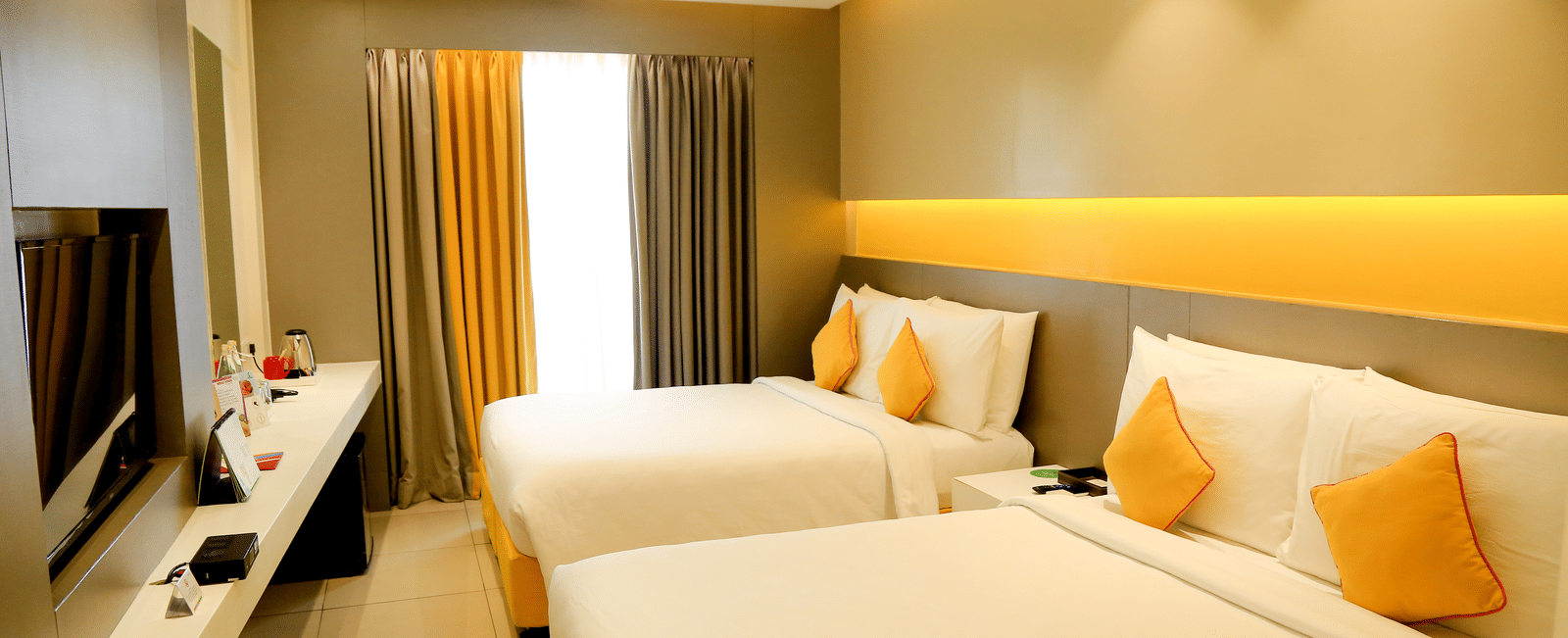 A spacious room with twin beds featuring yellow cushions and headboards, large windows with curtains, and a wall-mounted TV - Zibe Coimbatore by GRT Hotels