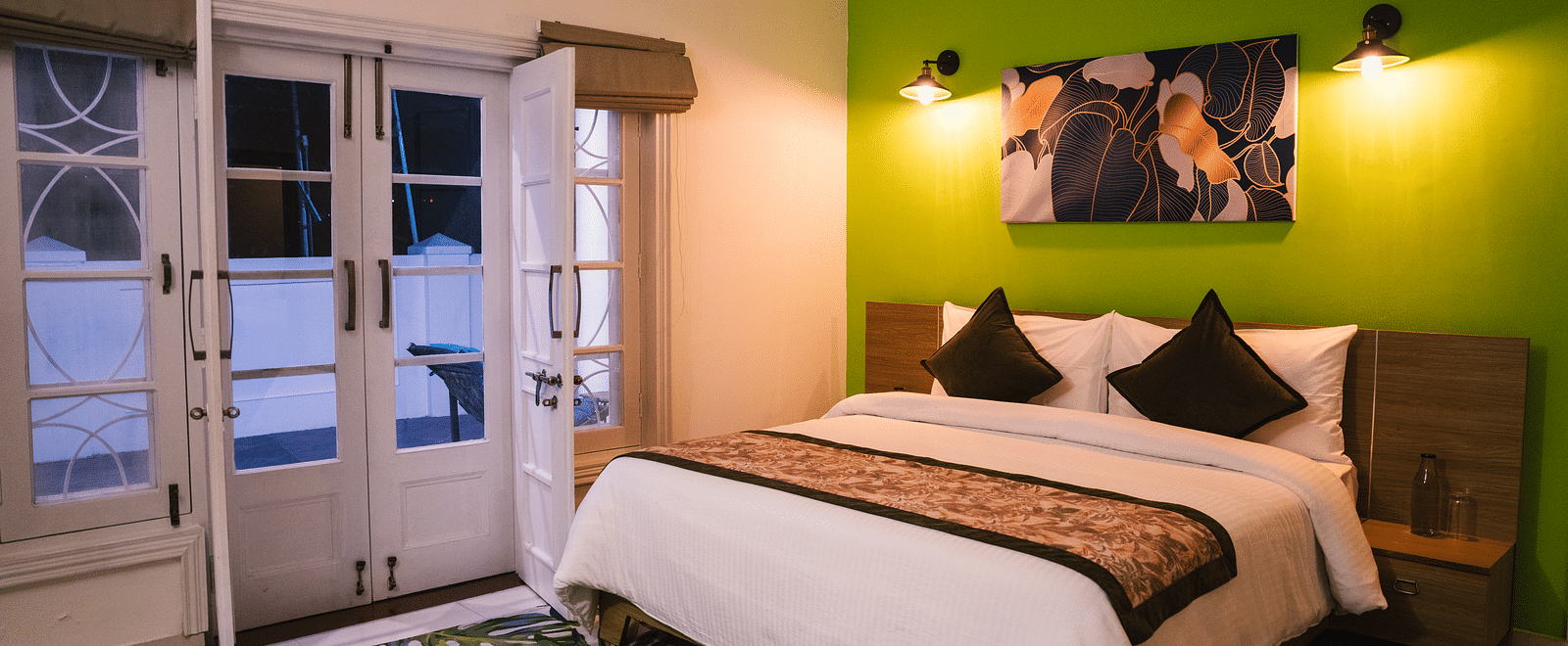 A spacious room at BluSalzz Villas - The Green Roof, Bhimtal, featuring a large bed with a photo frame above the headboard, night lamps glowing on either side, a carpet, and a door leading to the balcony.