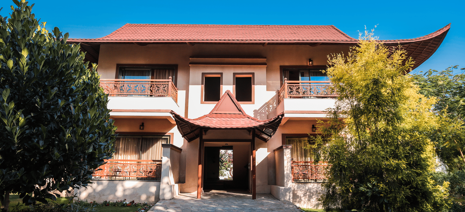 Two-storey building with Asian-style red tiled roof, decorative balconies, and a central entrance, surrounded by trees, plants, and a garden under a clear blue sky - Ananta Spa & Resort, Jaipur