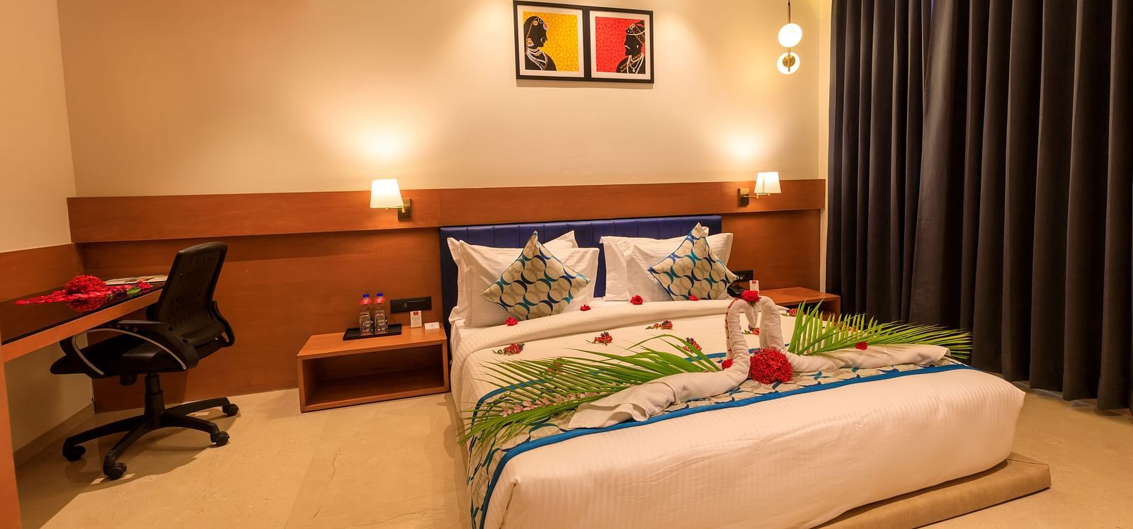 A warmly lit hotel room at Lords Eco Inn Apple Gate features a decorated double bed, bedside tables, a desk with a chair, and dark curtains.