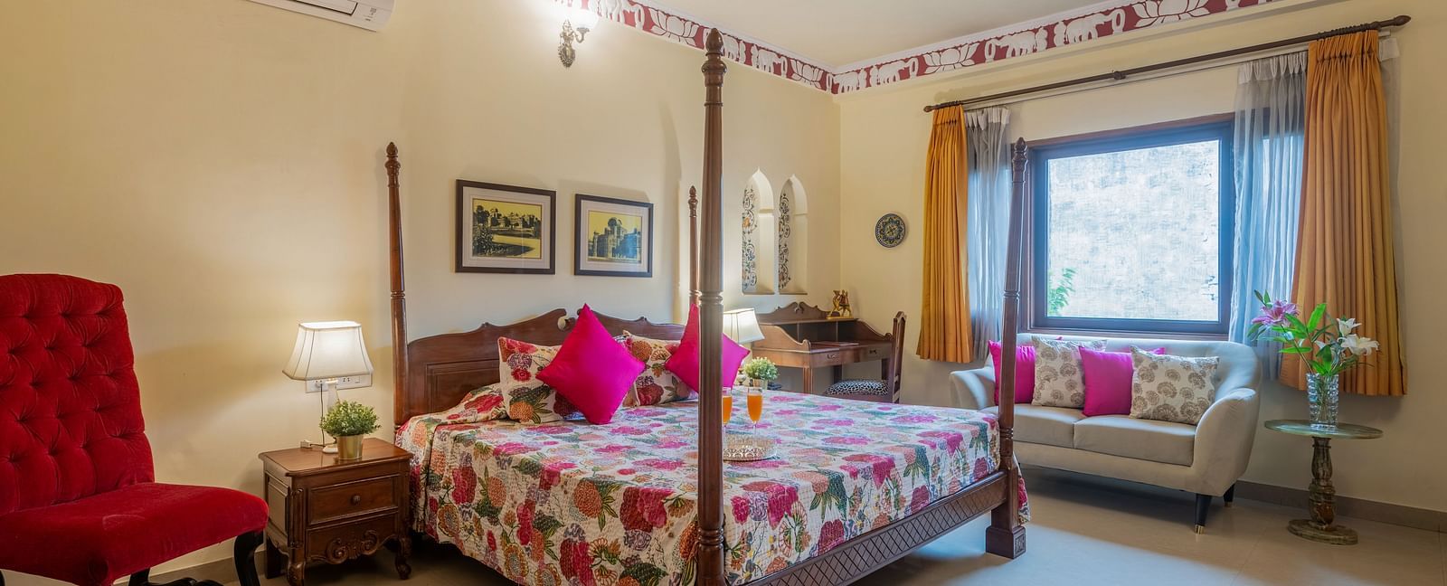 A bedroom with a four-poster bed, patterned bedding, pink cushions, a red armchair, and decorative trim along the ceiling | Dera Rawatsar, Jaipur