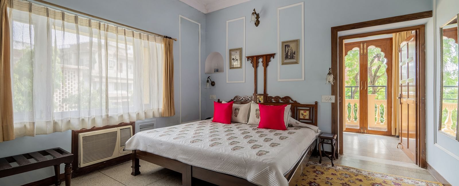 A bright and airy bedroom at Dera Rawatsar, Jaipur, features a comfortable bed, large windows with sheer curtains, and an ornate doorway leading outside.