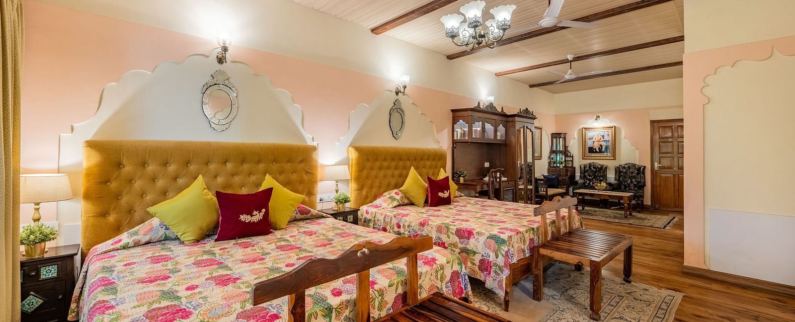 A grand suite at Dera Rawatsar, Jaipur, features two comfortable beds, a seating area with plush sofas, wooden beams, and elegant chandeliers.