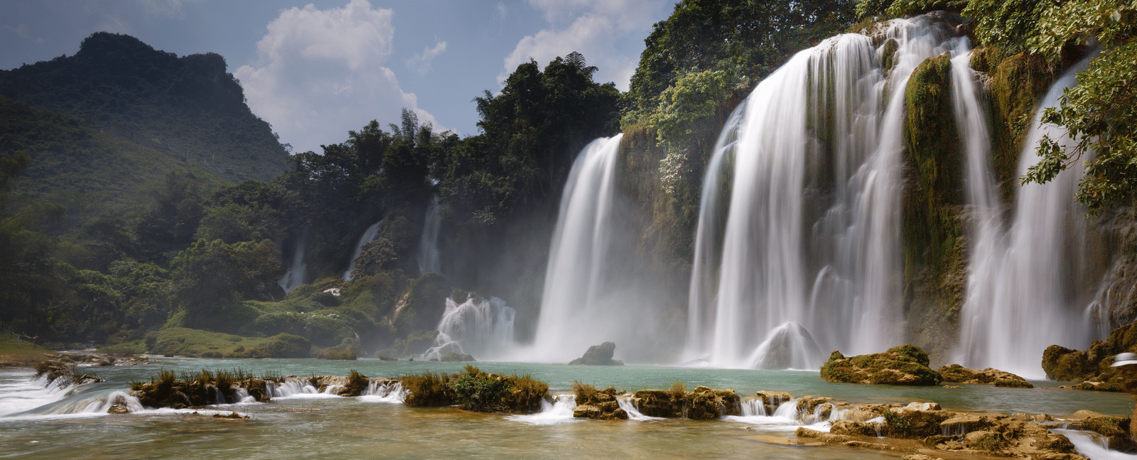 A wide cascade of water tumbles into a calm, wide river surrounded by lush trees on a sunny day