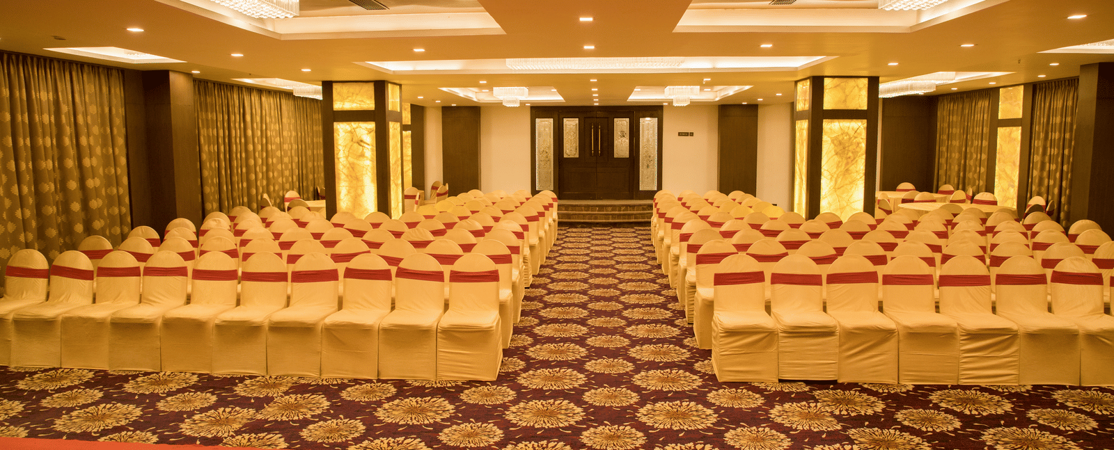 A banquet hall at Benzz Park Chennai arranged with numerous chairs in rows, featuring a patterned carpet and warm lighting.