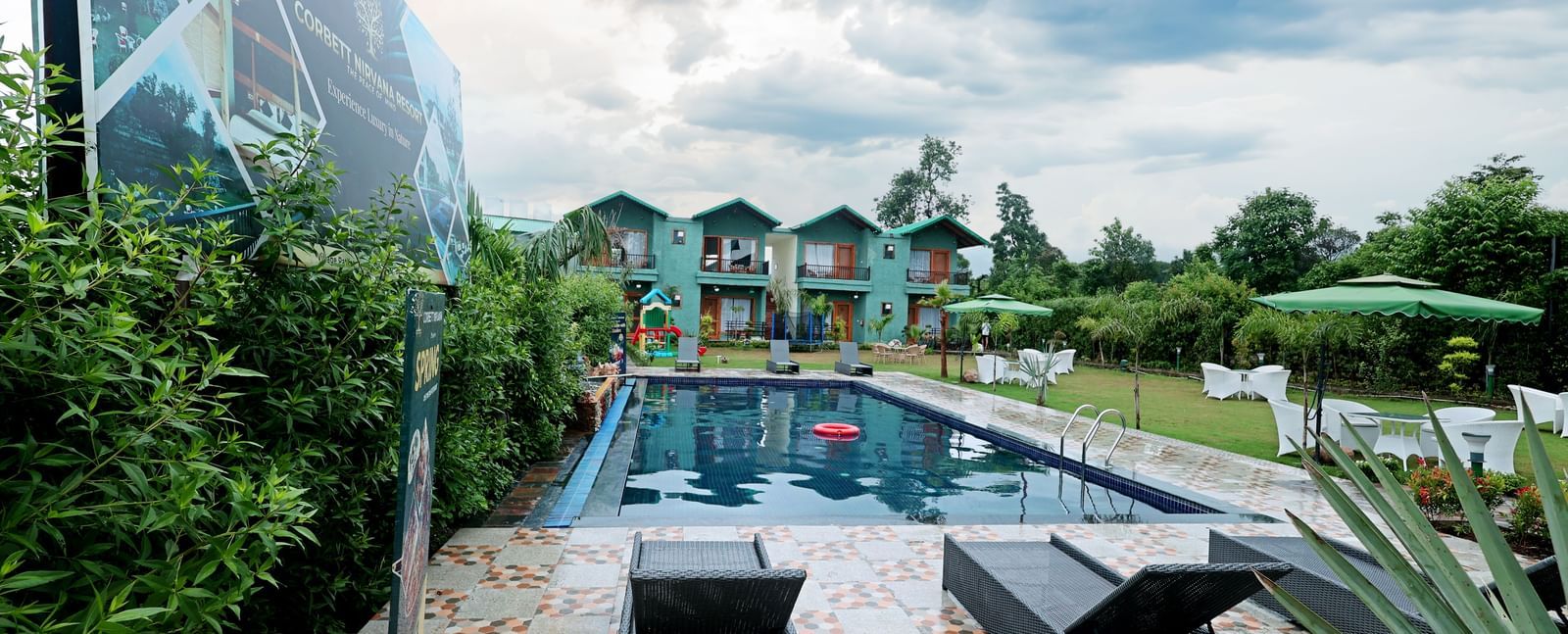 Guests can relax on sun loungers by the pool with a view of the modern resort buildings at Corbett Nirvana Resort.