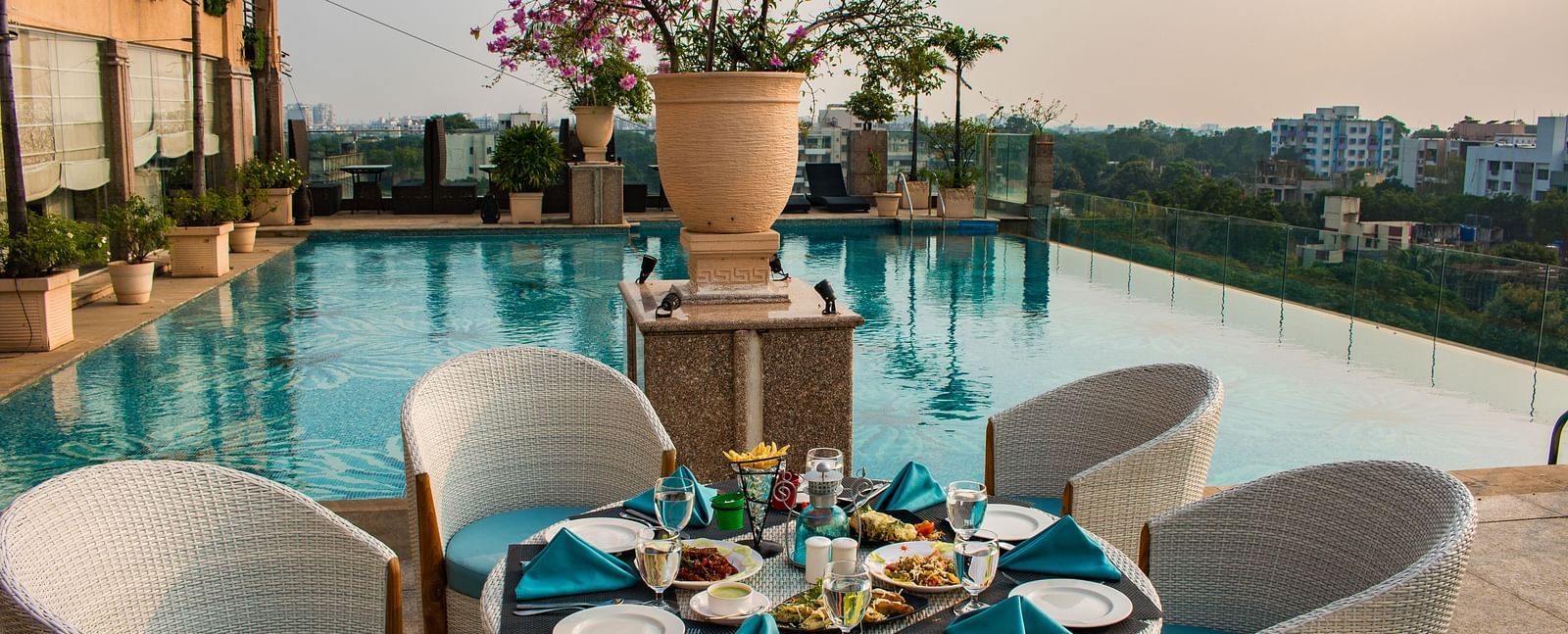 An outdoor dining at Hotel Ashok Nagpur featuring a round table with place settings for four, surrounded by cushioned chairs, and a swimming pool and cityscape in the background. 
