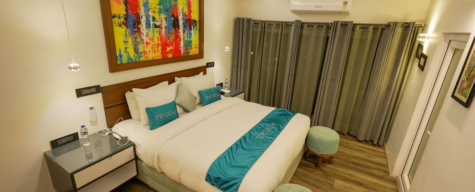 Bedroom of Pent House at The Tattva, Joshimath with a large bed, teal accents, abstract wall art, wooden flooring, grey curtains, and ambient ceiling lighting.