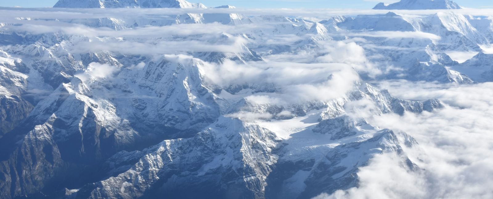 An aerial view of a vast range of snow-capped mountains and jagged peaks extending to the horizon, partially obscured by white clouds and mist under a clear blue sky.