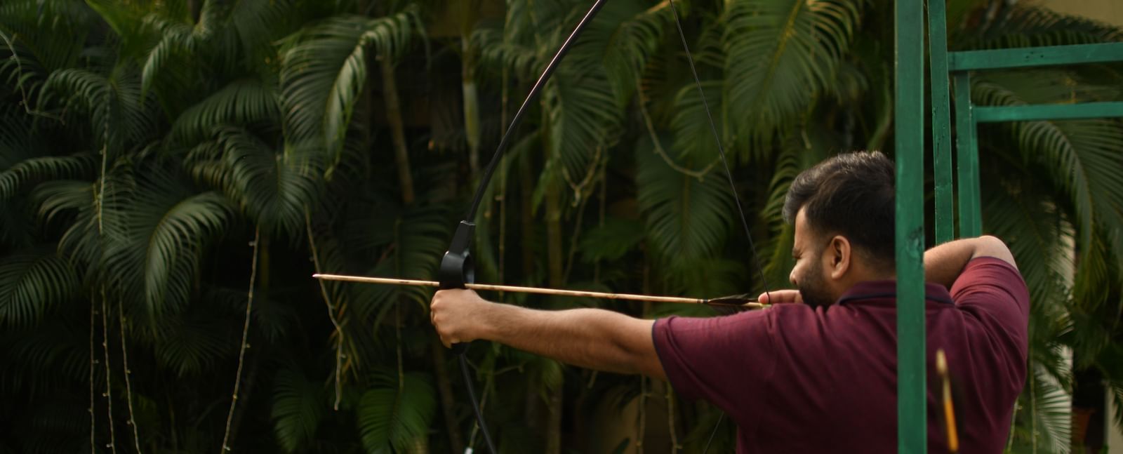 A person aims a long blowgun or similar weapon in a dense, green, natural environment