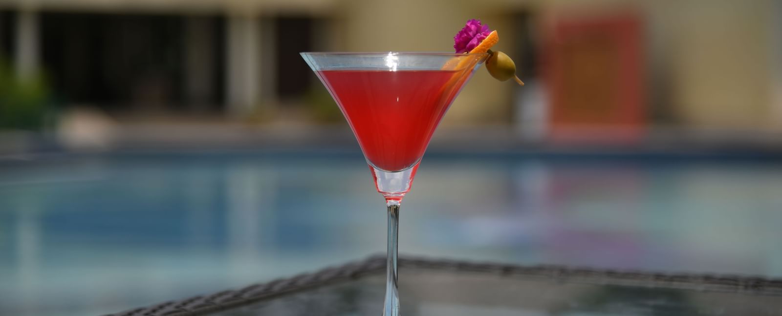 A red drink with a garnish in a glass by a swimming pool at Vedic Village Spa Resort.
