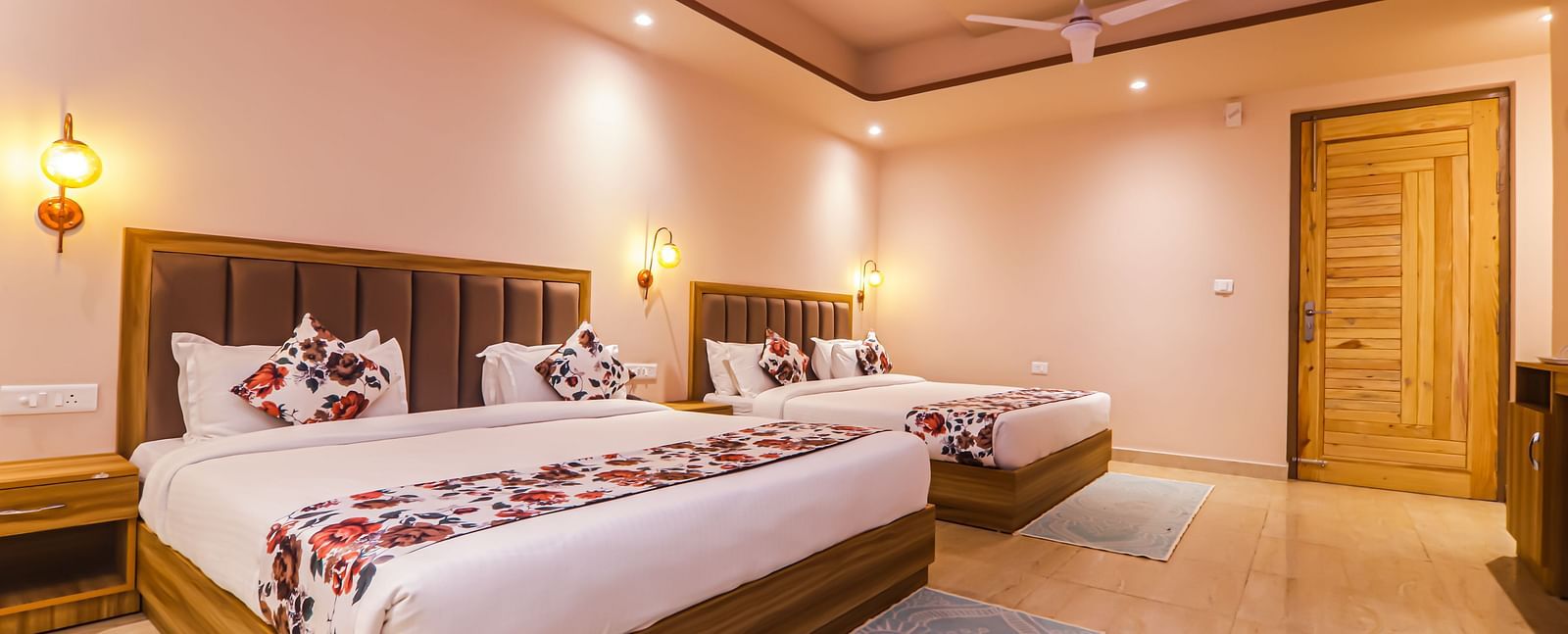A well-lit bedroom at Vedikant Resorts The Mallard Corbett features two beds with floral patterned runners and pillows, wooden frames, and a ceiling fan.