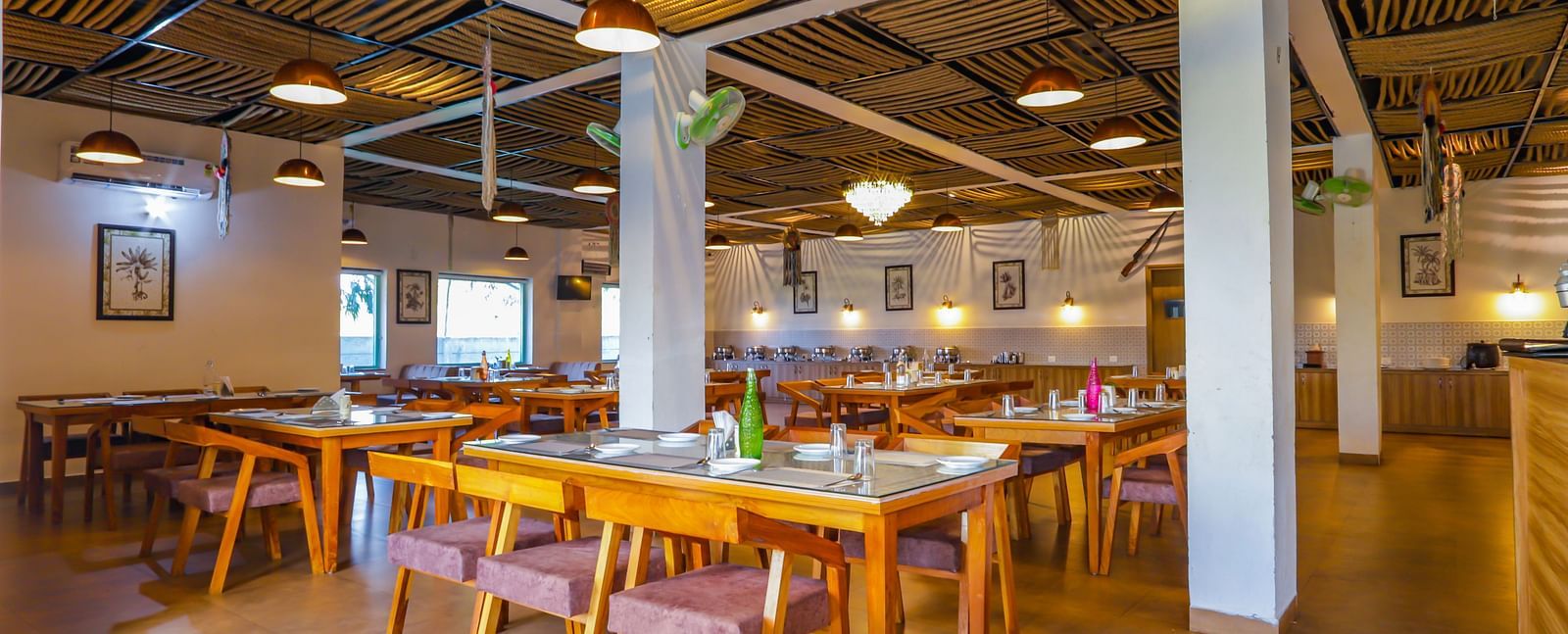 A spacious dining hall at Vedikant Resorts The Mallard Corbett features wooden tables and chairs under a high ceiling with lights.