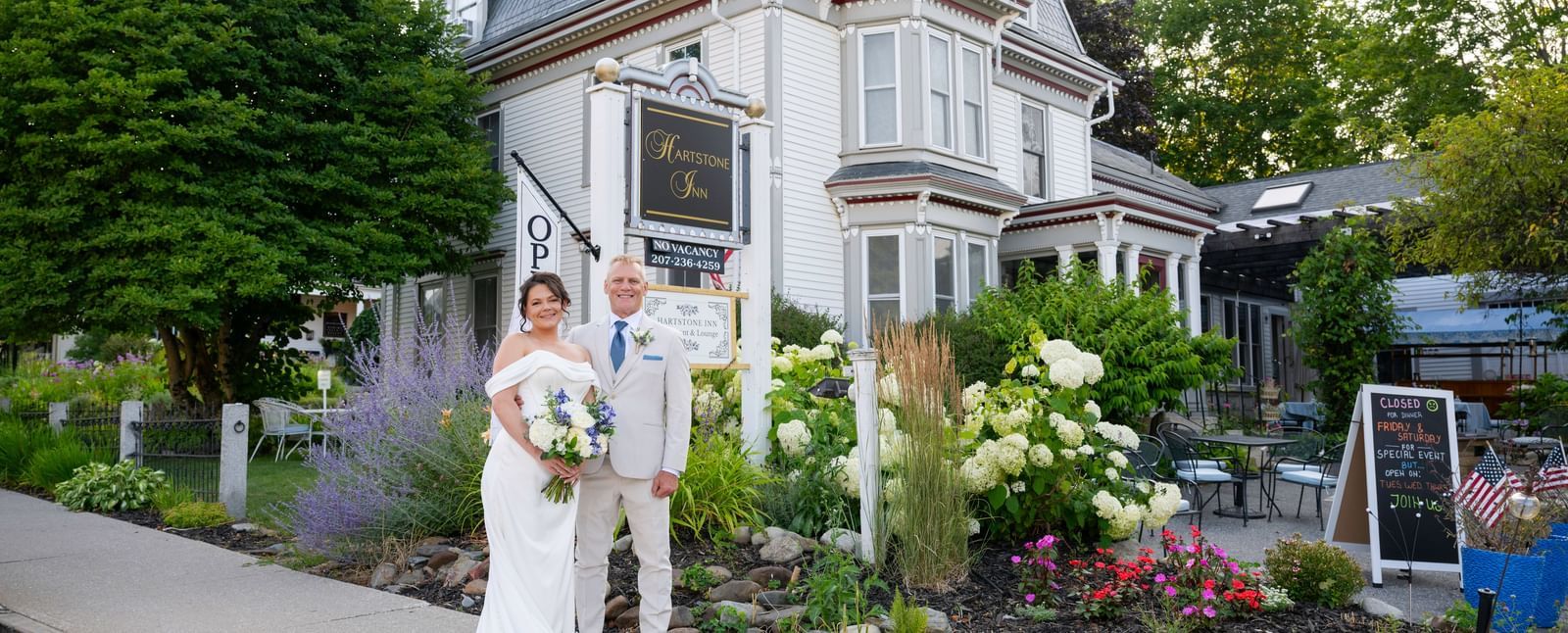 A bride in a dress and a groom in a suit stand in front of the Hartstone Inn sign, with flowers, a tree, and the building visible in the background.