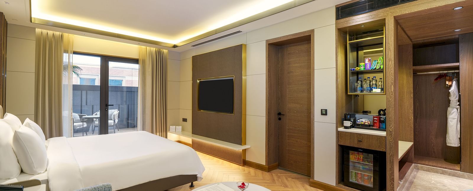 A modern hotel suite at Nirvana Luxury Hotel, Ludhiana, featuring a double bed, a plush blue settee, a round table with snacks, a wall-mounted telly, a minibar, and access to a balcony.
