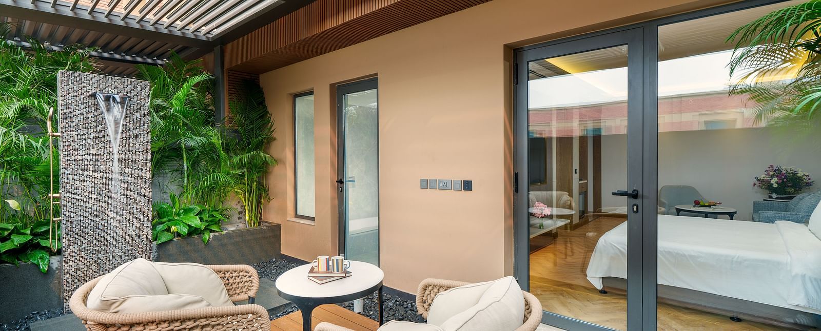 A luxurious villa patio at Nirvana Luxury Hotel, Ludhiana, with comfy outdoor armchairs, a small round table, a stylish water feature, lush plants, and a glimpse into the villa's bedroom.