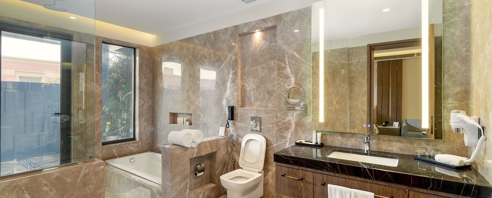A luxurious bathroom at Nirvana Luxury Hotel, Ludhiana, showcases marble walls and flooring, a white bathtub by a window, a modern toilet, and a dark countertop with a sink and mirror.