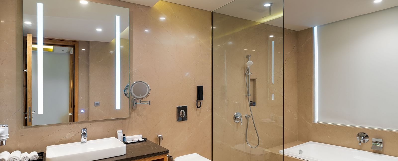 A well-appointed hotel bathroom at Nirvana Luxury Hotel, Ludhiana, featuring a rectangular sink with a large, illuminated mirror, a toilet, and a separate glass-enclosed shower and bathtub.