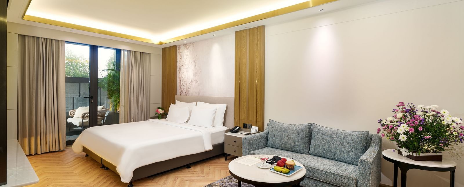 A stylish hotel room at Nirvana Luxury Hotel, Ludhiana, with a comfy bed, a smart blue sofa, a round table with fruit, and a colourful rug on a herringbone floor.