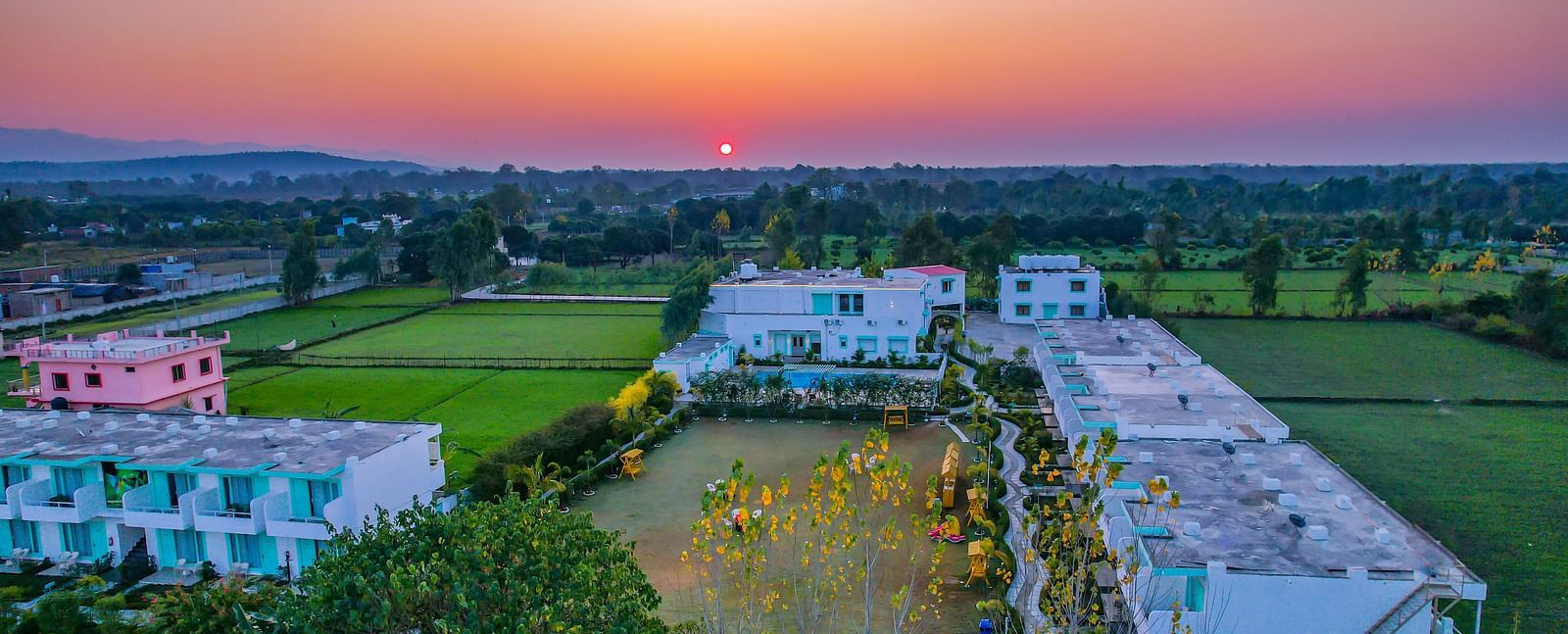 An aerial view of Vedikant Resorts The Mallard Corbett at sunset shows buildings and surrounding greenery.